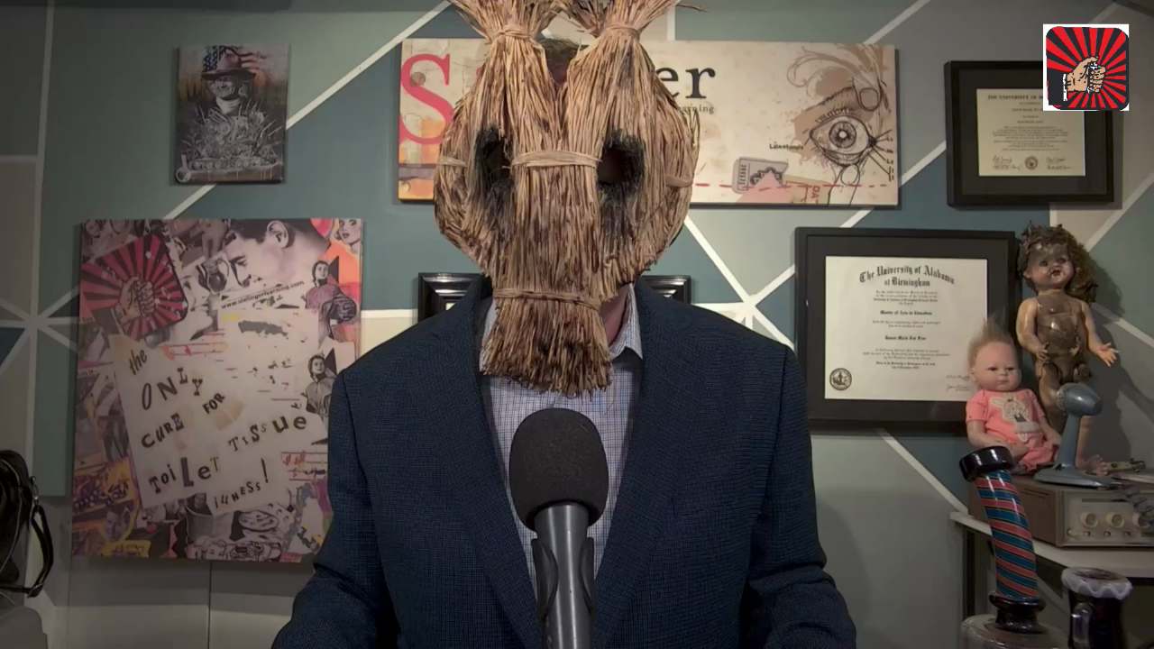 SFL podcast host wearing a straw mask in front of collage art, framed diplomas, dolls, and a microphone.